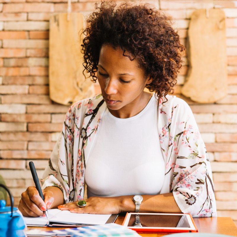 young-woman-writing-diary-with-pen-digital-tablet-wooden-table